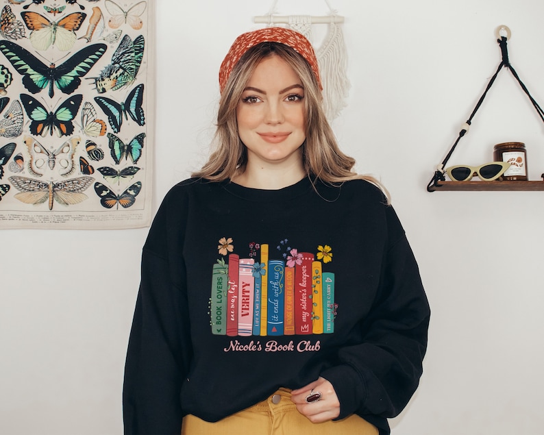 Op de afbeelding: Zwart sweatshirt met een afbeelding van kleurrijke boeken met bloemmotieven en de tekst "Nicole's Book Club".