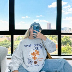 May include: A gray sweatshirt with a graphic print of a plate of croissants and a cup of coffee. The text on the sweatshirt reads "Sunday Brunch Club" and "Every Sunday at 11 AM" and "Un croissant et un cafe".