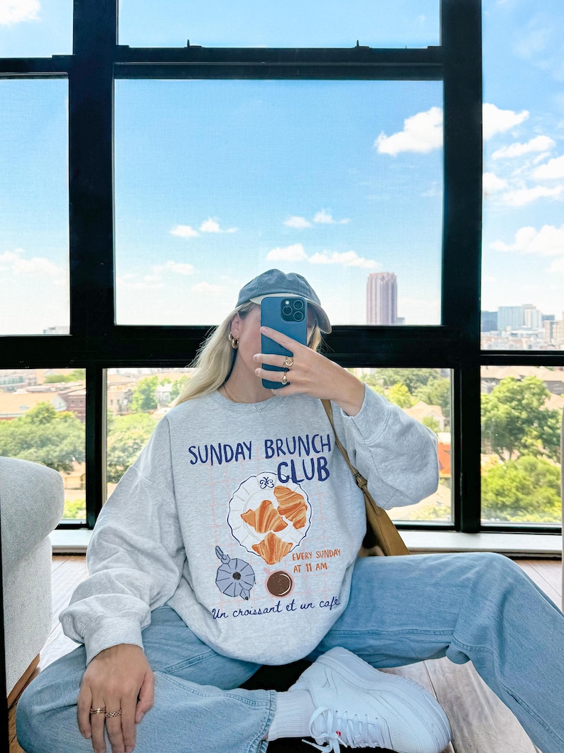 May include: A gray sweatshirt with a graphic print of a plate of croissants and a cup of coffee. The text on the sweatshirt reads "Sunday Brunch Club" and "Every Sunday at 11 AM" and "Un croissant et un cafe".