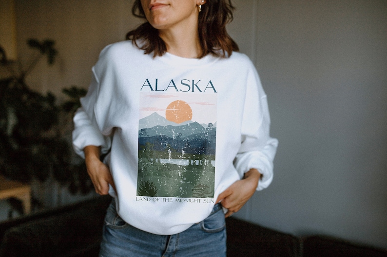 Pu&ograve; includere: Felpa bianca con una stampa grafica di un paesaggio montano e il testo "Alaska, Land of the Midnight Sun".
