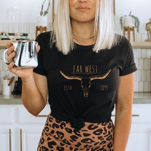 K&ouml;nnte beinhalten: Eine Frau tr&auml;gt ein schwarzes T-Shirt mit einer goldenen Grafik eines Longhorn-Sch&auml;dels und dem Text "Far West Estd 1899". Sie h&auml;lt eine schwarz-wei&szlig;e marmorierte Tasse.