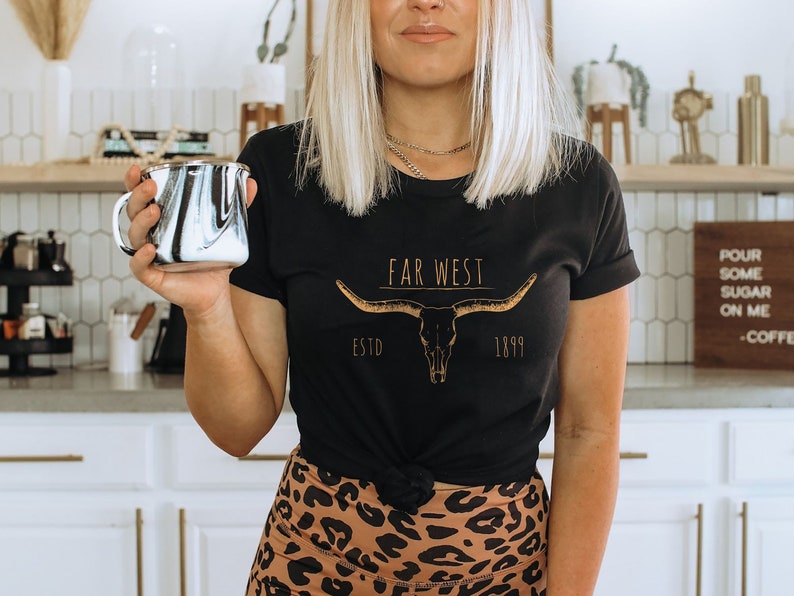 K&ouml;nnte beinhalten: Eine Frau tr&auml;gt ein schwarzes T-Shirt mit einer goldenen Grafik eines Longhorn-Sch&auml;dels und dem Text "Far West Estd 1899". Sie h&auml;lt eine schwarz-wei&szlig;e marmorierte Tasse.