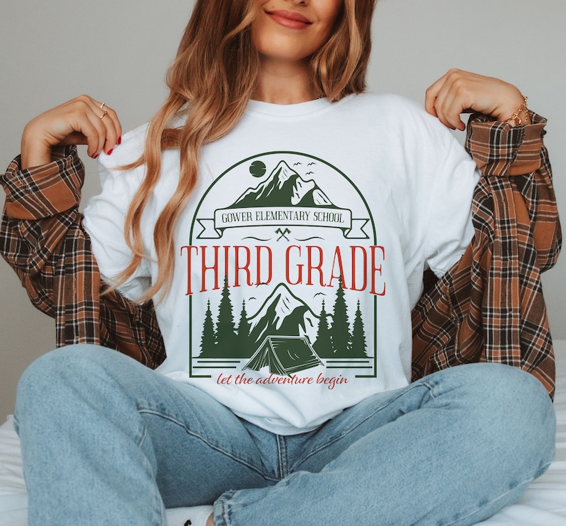 May include: White t-shirt featuring a green and brown mountain scene graphic. The graphic includes a tent, trees, and the text "Gower Elementary School Third Grade Let the adventure begin".