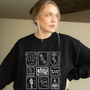 Op de afbeelding: Zwart sweatshirt met een witte grafische print van een reeks postzegels. De postzegels bevatten verschillende ontwerpen, waaronder een hand, een roos, een huis, een vogel en een hart. De tekst op de postzegels is "Minus Finn Pyri", "Like a rose in a graveyard", "Be careful. Don't die" en "What's next?"