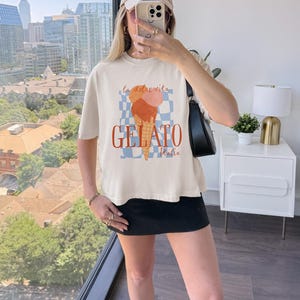 May include: Cream-colored t-shirt featuring a gelato cone graphic with the words "la dolce vita" and "GELATO Italia" in red. The design includes a blue and white checkered background. The shirt is worn by a person.