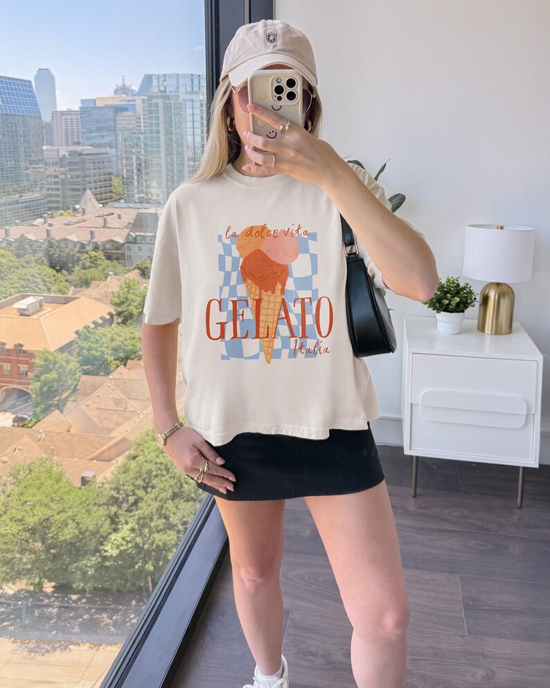May include: Cream-colored t-shirt featuring a gelato cone graphic with the words "la dolce vita" and "GELATO Italia" in red. The design includes a blue and white checkered background. The shirt is worn by a person.