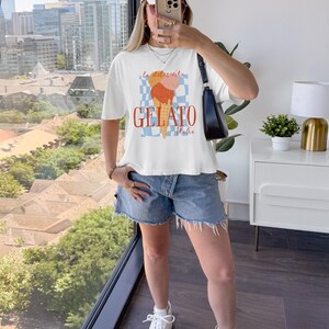 May include: White t-shirt with a gelato graphic. The design features an ice cream cone with orange and pink scoops, the word "GELATO" in orange, and the text "la dolce vita" and "Italia". The model is wearing a white cap and denim shorts.