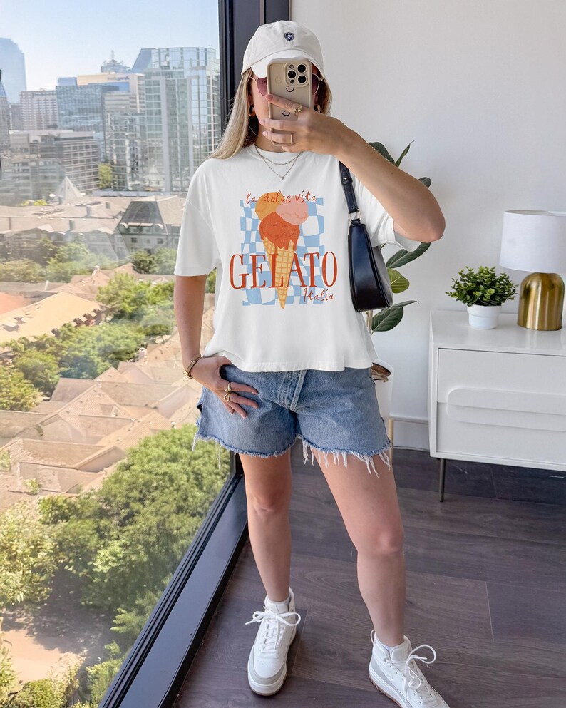 May include: White t-shirt with a gelato graphic. The design features an ice cream cone with orange and pink scoops, the word "GELATO" in orange, and the text "la dolce vita" and "Italia". The model is wearing a white cap and denim shorts.