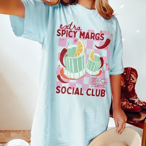 May include: A light blue t-shirt with a graphic of two margarita glasses with a lime wedge and chili peppers. The text "extra spicy margs social club" is printed on the shirt.