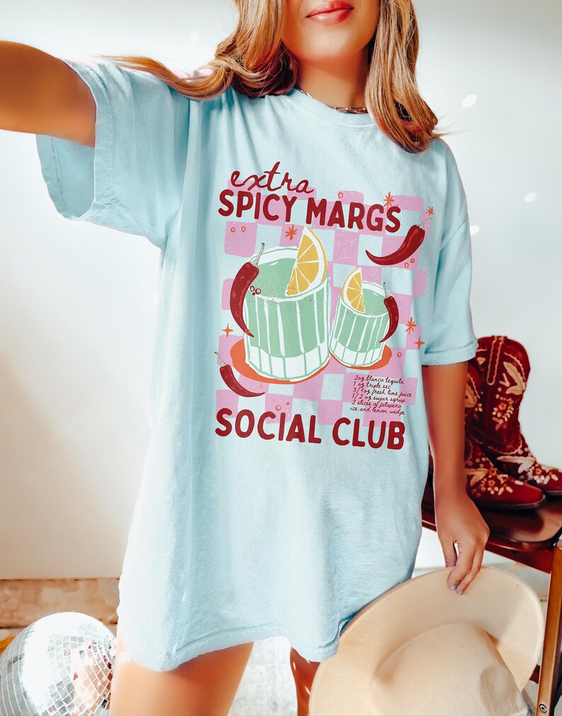 May include: A light blue t-shirt with a graphic of two margarita glasses with a lime wedge and chili peppers. The text "extra spicy margs social club" is printed on the shirt.