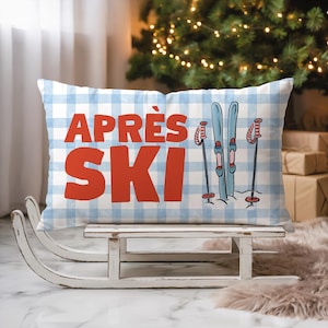 Può includere: Un cuscino rettangolare con un motivo a quadri blu e bianchi. Il cuscino presenta le parole "APRÈS SKI" in lettere rosse, insieme a una grafica di sci e bastoncini. Il cuscino è appoggiato su una slitta di legno bianca.