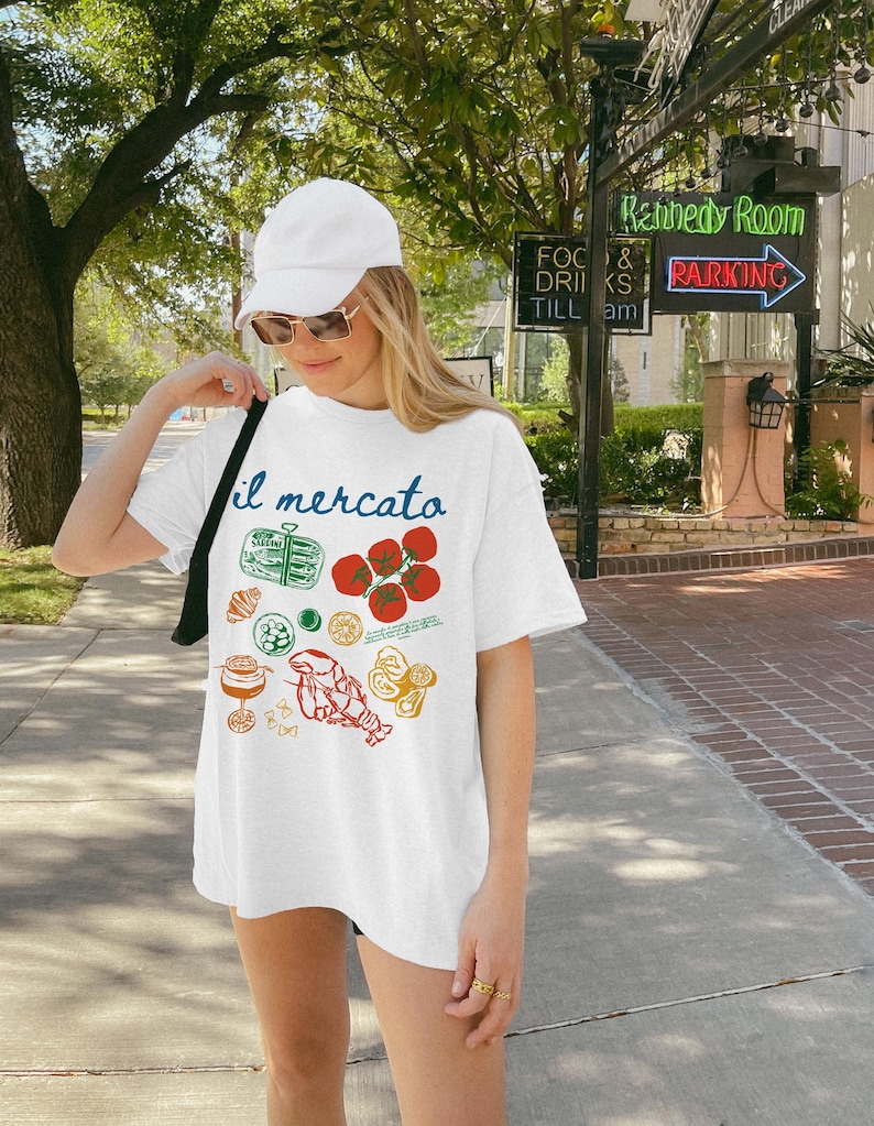 May include: White t-shirt with a graphic print of various Italian food items, including tomatoes, pasta, and a lobster. The text "il mercato" is printed on the shirt.