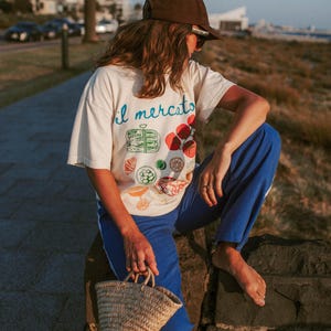 May include: A person wearing a white t-shirt with the text "il mercato" and a colorful illustration of food. They are wearing blue pants and a brown baseball cap. They are sitting on a stone wall and holding a small woven basket.