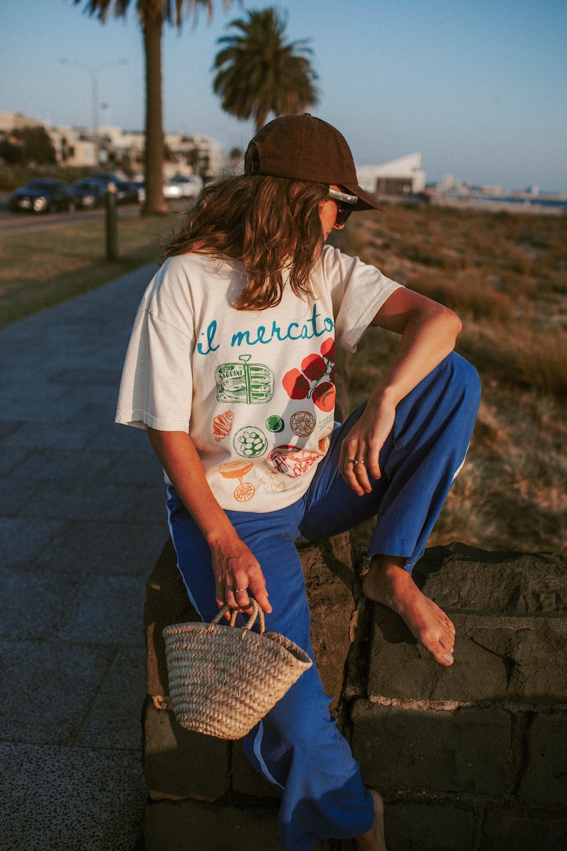 May include: A person wearing a white t-shirt with the text "il mercato" and a colorful illustration of food. They are wearing blue pants and a brown baseball cap. They are sitting on a stone wall and holding a small woven basket.