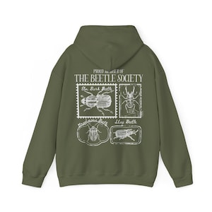 K&ouml;nnte beinhalten: Ein gr&uuml;ner Kapuzenpullover mit einem wei&szlig;en Grafikdruck einer Sammlung von Vintage-Briefmarken mit verschiedenen K&auml;ferarten. Der Text auf den Briefmarken lautet "Elm Bark Beetle", "Stag Beetle" und "Strabosoma Beetle". Der Text &uuml;ber den Briefmarken lautet "Proud member of The Beetle Society".