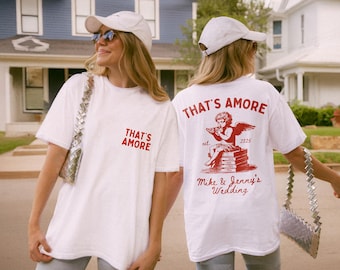 Op de afbeelding: Twee witte T-shirts met rode tekst. Eén shirt zegt "That's Amore" met een kleine illustratie van een cupido die een boek vasthoudt. Het andere shirt zegt "That's Amore" met de tekst "Mike & Jenny's Wedding" eronder.