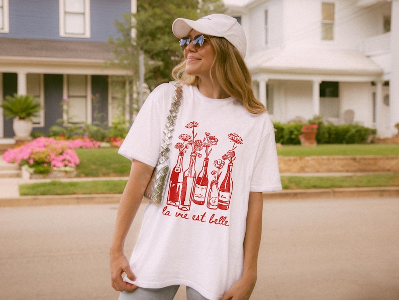 Peut inclure: T-shirt blanc avec un motif rouge de bouteilles de vin avec des fleurs &agrave; l'int&eacute;rieur. Le texte "la vie est belle" est imprim&eacute; sous le motif.