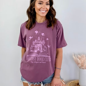 May include: A purple t-shirt with a graphic of a castle, book, and dragons. The text on the shirt reads "Fantasy Book Club: magic, dragons and fairies".