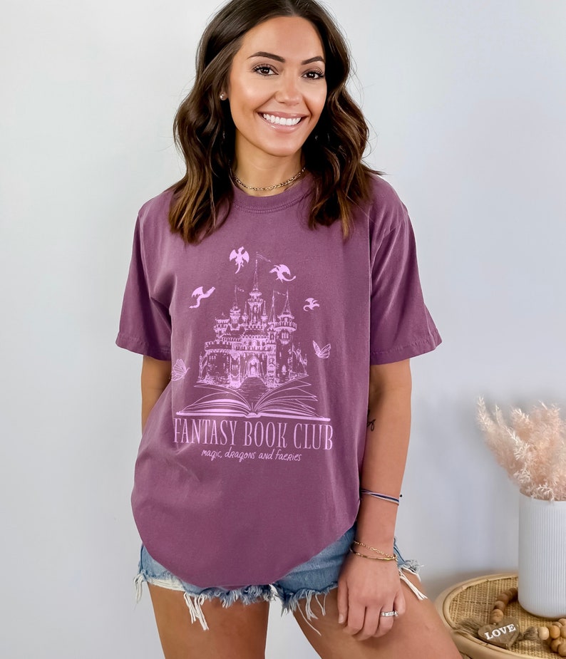 May include: A purple t-shirt with a graphic of a castle, book, and dragons. The text on the shirt reads "Fantasy Book Club: magic, dragons and fairies".