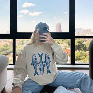 May include: A person wearing a beige sweatshirt with a blue fish graphic, blue jeans, and a blue baseball cap. The person is holding a cell phone in front of their face.