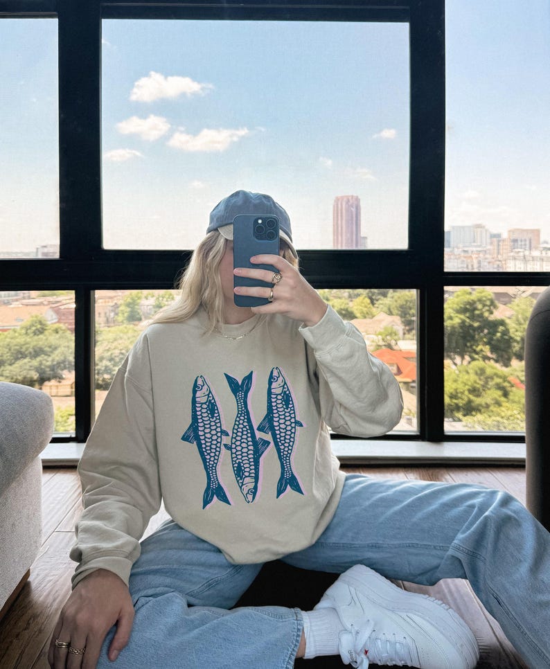 May include: A person wearing a beige sweatshirt with a blue fish graphic, blue jeans, and a blue baseball cap. The person is holding a cell phone in front of their face.