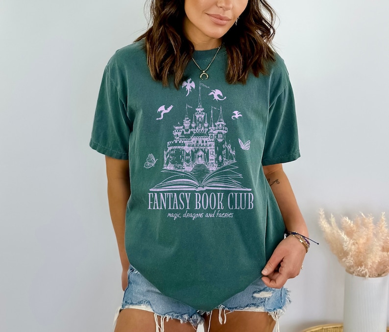 May include: A dark green t-shirt with a graphic of a castle, dragons, and a book. The text "Fantasy Book Club" is printed below the graphic, with the words "magic, dragons and fairies" printed below that.