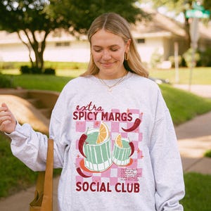 May include: A light grey sweatshirt with the text "extra SPICY MARGS SOCIAL CLUB" in pink and red. The graphic features two margarita glasses with orange slices and chilli peppers on a pink checkered background.