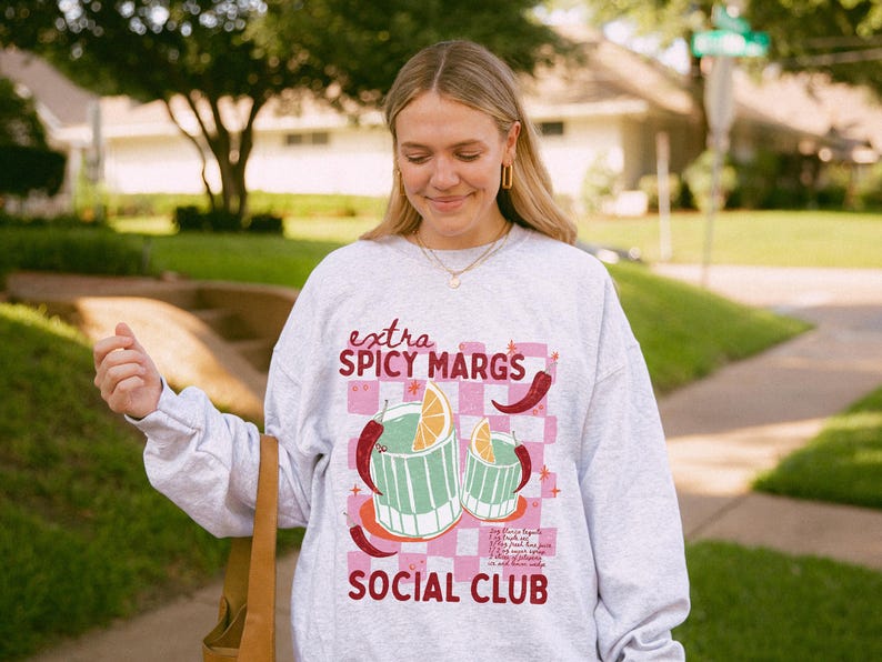 May include: A light grey sweatshirt with the text "extra SPICY MARGS SOCIAL CLUB" in pink and red. The graphic features two margarita glasses with orange slices and chilli peppers on a pink checkered background.