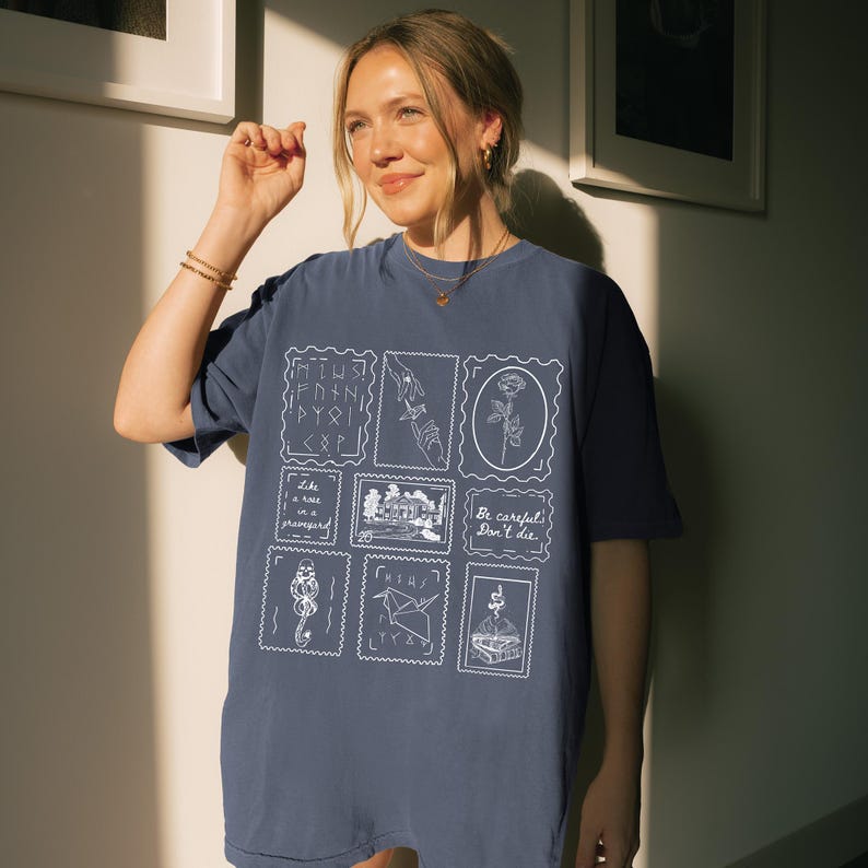 May include: A navy blue t-shirt featuring a collection of white illustrations within postage stamp-like frames. The designs include a rose, hands, a house, and text that reads "Be careful, Don't die." The shirt has a relaxed fit.