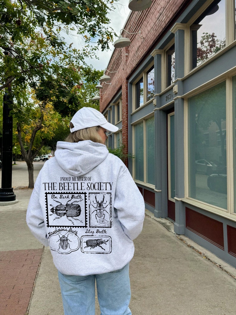 K&ouml;nnte beinhalten: Ein grauer Hoodie mit einem schwarzen Aufdruck, der "Proud Member of The Beetle Society" lautet und vier verschiedene K&auml;ferarten zeigt. Der Hoodie hat einen wei&szlig;en Kordelzug und eine wei&szlig;e Baseballkappe wird auf dem Kopf der Person getragen.