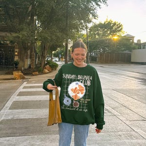 May include: A woman wearing a green sweatshirt with the text "Sunday Brunch Club" and a graphic of croissants, coffee, and a plate. She is also wearing blue jeans and carrying a brown tote bag.