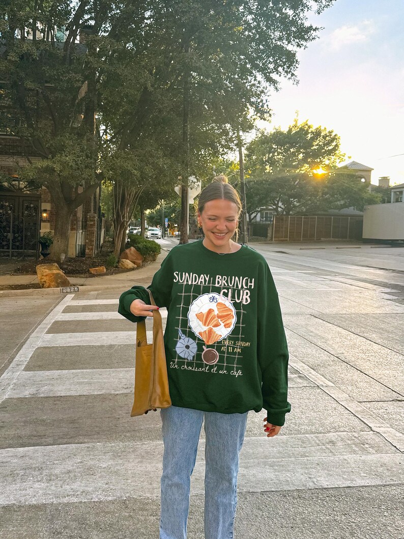 May include: A woman wearing a green sweatshirt with the text "Sunday Brunch Club" and a graphic of croissants, coffee, and a plate. She is also wearing blue jeans and carrying a brown tote bag.