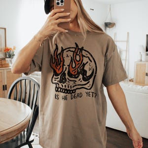 May include: A brown oversized t-shirt with a graphic of a skull with flames for eyes. The text "Is he dead yet?" is printed below the skull.