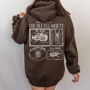 K&ouml;nnte beinhalten: Ein brauner Hoodie mit einem wei&szlig;en Grafikdruck, auf dem "Proud Member of The Beetle Society" steht. Die Grafik zeigt Illustrationen verschiedener K&auml;ferarten, darunter ein Ulmenrindenk&auml;fer, ein Hirschk&auml;fer und ein Graphosoma-K&auml;fer.