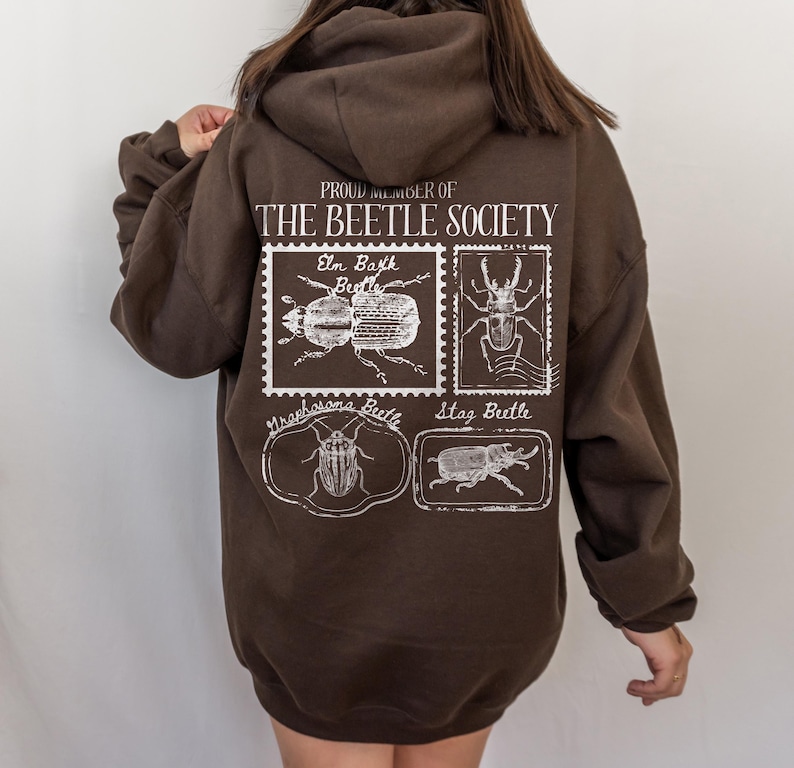 K&ouml;nnte beinhalten: Ein brauner Hoodie mit einem wei&szlig;en Grafikdruck, auf dem "Proud Member of The Beetle Society" steht. Die Grafik zeigt Illustrationen verschiedener K&auml;ferarten, darunter ein Ulmenrindenk&auml;fer, ein Hirschk&auml;fer und ein Graphosoma-K&auml;fer.