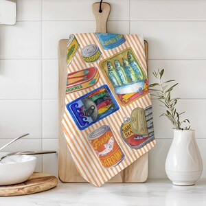 May include: A kitchen tea towel with a striped orange and white background. The towel features a repeating pattern of colourful illustrations of canned sardines. The text on the cans reads "Canned Fish", "Sardines", "Fresh Sardines", and "Sardines Sea".