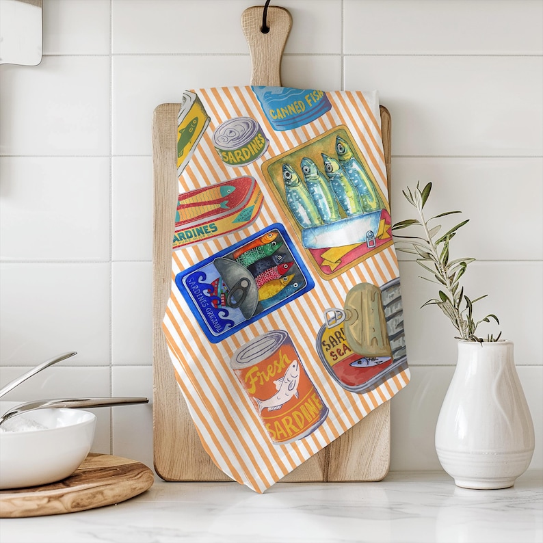 May include: A kitchen tea towel with a striped orange and white background. The towel features a repeating pattern of colourful illustrations of canned sardines. The text on the cans reads "Canned Fish", "Sardines", "Fresh Sardines", and "Sardines Sea".
