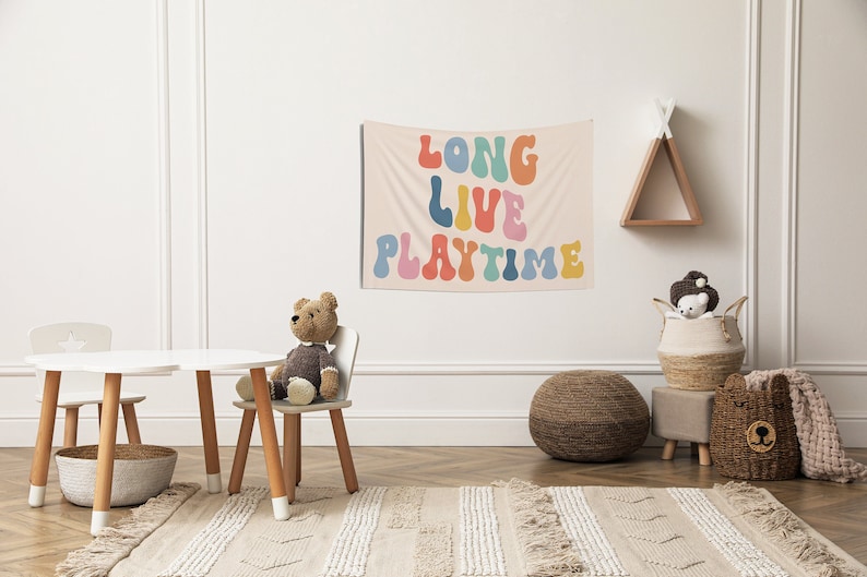 May include: A white wall with a colourful tapestry that reads "Long Live Playtime" in a retro font. A brown teddy bear sits in a white wooden chair. There is a white wooden table with two chairs, a brown woven ottoman, and a brown woven basket with a bear face on it.