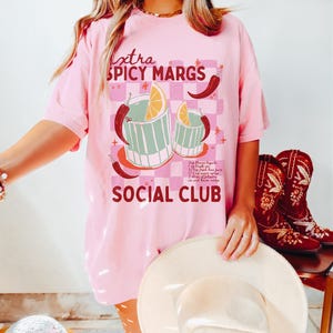 May include: Pink t-shirt with a graphic of two margarita glasses with a lime wedge and chili peppers. The text "extra spicy margs social club" is printed on the shirt.