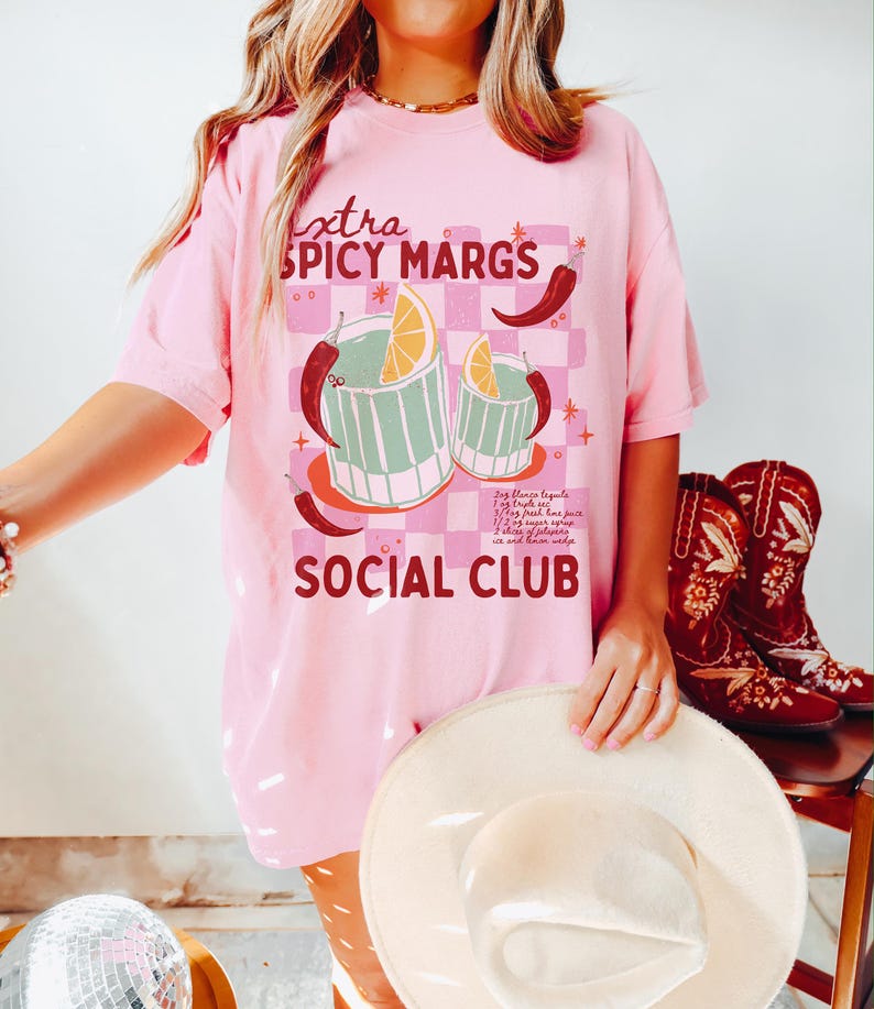 May include: Pink t-shirt with a graphic of two margarita glasses with a lime wedge and chili peppers. The text "extra spicy margs social club" is printed on the shirt.