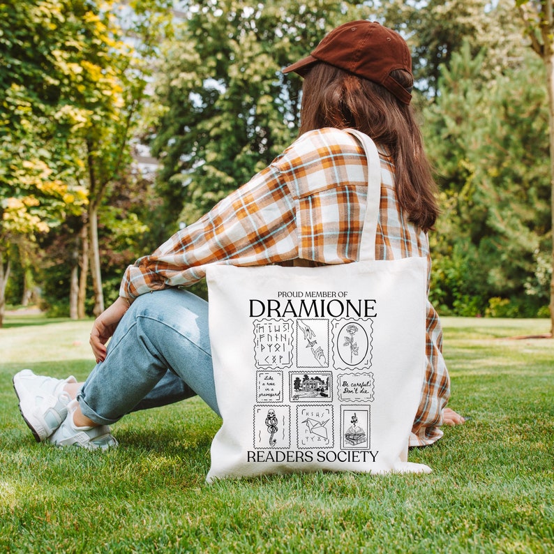 May include: White canvas tote bag with black text and illustrations. The text reads "Proud Member of Dramione Readers Society". The illustrations are of stamps with various Harry Potter themed images.
