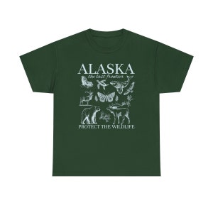 Puede incluir: Una camiseta verde oscuro con un dise&ntilde;o gr&aacute;fico blanco que presenta el texto "Alaska the last frontier" e ilustraciones de diversas especies de vida silvestre de Alaska, incluyendo un oso, una ballena, un alce, una mariposa y un p&aacute;jaro. El texto "Protect the Wildlife" est&aacute; impreso debajo de las ilustraciones.