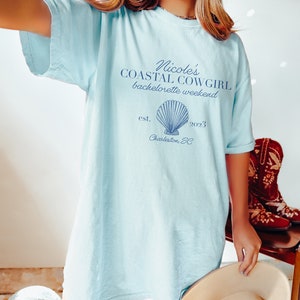 K&ouml;nnte beinhalten: Hellblaues T-Shirt mit dem Text "Nicole's Coastal Cowgirl bachelorette weekend est. 2023 Charleston, SC" und einer blauen Muschelgrafik.