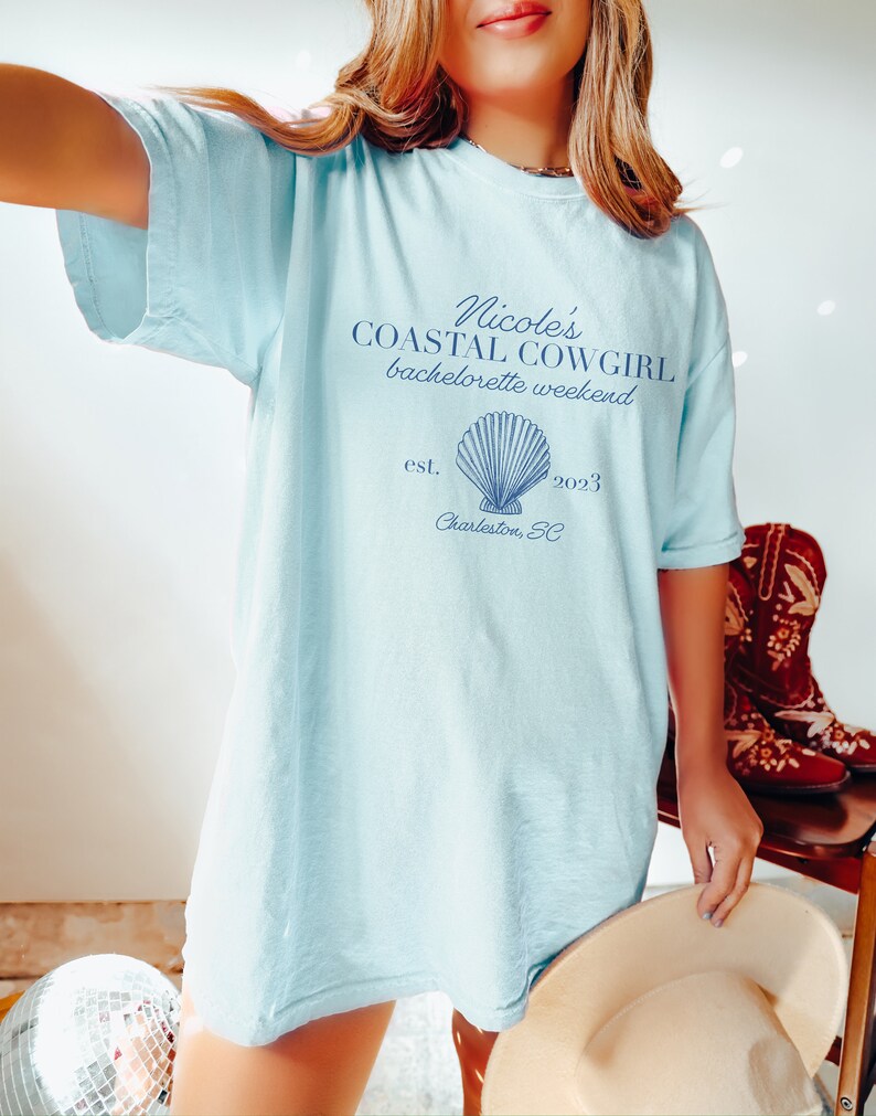 K&ouml;nnte beinhalten: Hellblaues T-Shirt mit dem Text "Nicole's Coastal Cowgirl bachelorette weekend est. 2023 Charleston, SC" und einer blauen Muschelgrafik.
