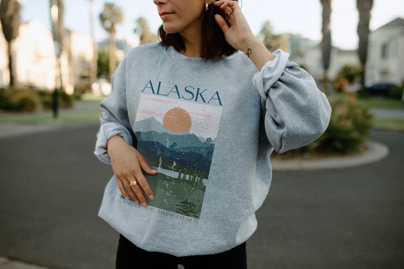 Pu&ograve; includere: Felpa grigia con una stampa grafica di una catena montuosa e un sole al tramonto. Il testo "Alaska" e "Land of the Midnight Sun" &egrave; stampato sulla felpa.