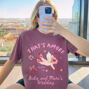 K&ouml;nnte beinhalten: Ein maroonfarbenes T-Shirt mit einem Grafikdruck eines Cupidos, der Cowboystiefel tr&auml;gt und einen Bogen und Pfeil h&auml;lt. Der Text "That's Amore" befindet sich &uuml;ber dem Cupido und "Katie and Marc's Wedding" unter dem Cupido. Der Text "est. 2025" befindet sich ebenfalls auf dem T-Shirt.