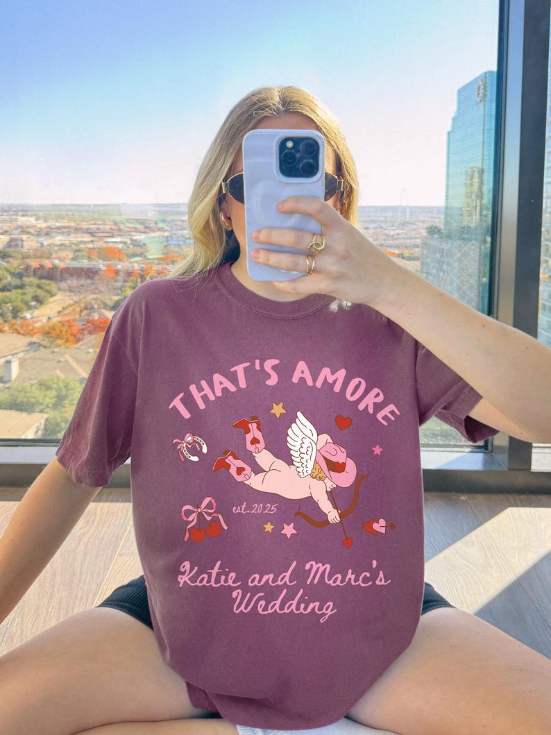 K&ouml;nnte beinhalten: Ein maroonfarbenes T-Shirt mit einem Grafikdruck eines Cupidos, der Cowboystiefel tr&auml;gt und einen Bogen und Pfeil h&auml;lt. Der Text "That's Amore" befindet sich &uuml;ber dem Cupido und "Katie and Marc's Wedding" unter dem Cupido. Der Text "est. 2025" befindet sich ebenfalls auf dem T-Shirt.