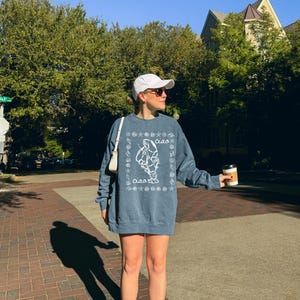 Pu&ograve; includere: Una donna indossa una felpa blu con una grafica bianca di una creatura marina e la parola "ciao". Indossa un cappello da baseball bianco e scarpe da ginnastica bianche. Tiene una tazza di caff&egrave; nella mano destra.