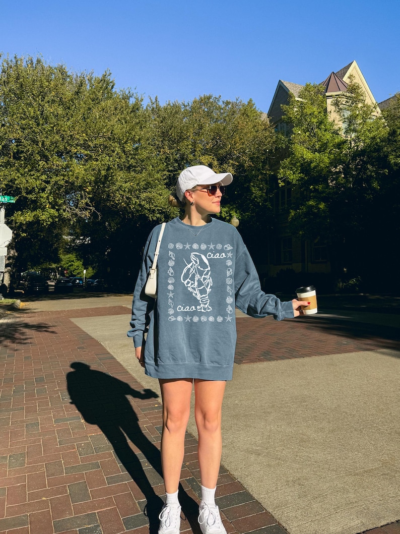 Pu&ograve; includere: Una donna indossa una felpa blu con una grafica bianca di una creatura marina e la parola "ciao". Indossa un cappello da baseball bianco e scarpe da ginnastica bianche. Tiene una tazza di caff&egrave; nella mano destra.
