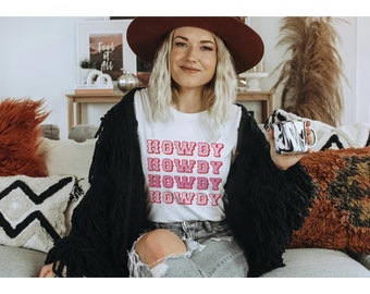 May include: A woman wearing a white t-shirt with the word "HOWDY" repeated four times in pink and red letters. She is wearing a black fringed cardigan and a brown hat. She is sitting on a sofa with a mug in her hand.
